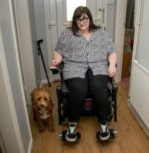 Woman in powerchair wearing heart print shirt, black jeans and trainers, next to her on wooden flooring is a red and white cockapoo