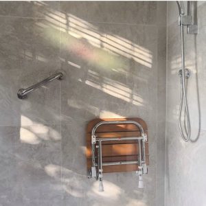 Grey tiled shower with chrome grab rails and a wooden shower chair folded up onto the wall