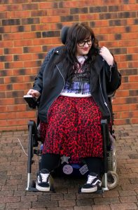 Plus size woman in wheelchair user wearing a black leather jacket, Stranger Things t-shirt, red and black animal print skirt, black tights and Vans