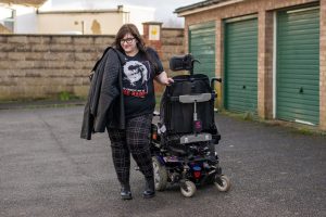 Woman stood next to wheelchair wearing Bowie from Labyrinth tshirt, red and black check trousers, black Dr Marten boots and holding a leather jacket over shoulder