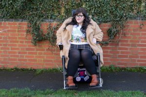 Plus size woman sat in wheelchair wearing brown mini skirt, black tights, Wizard of Oz t-shirt and cream teddy coat