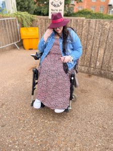 Plus size white woman sat in wheelchair wearing maxi floral dress, denim jacket, trainers and burgundy fedora hat